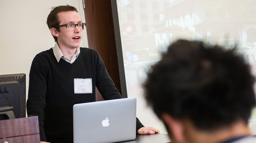 A person with glasses, wearing a black sweater over a white shirt, is speaking at a lectern with an open laptop in front. An audience member is visible in the foreground. A screen with a presentation is visible in the background.